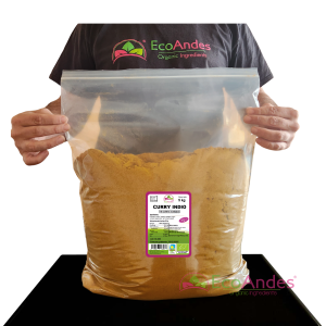 Persona con camiseta de la marca EcoAndes sosteniendo una bolsa transparente con cierre zip de 5 kg de Curry indio en polvo. El empaque muestra una etiqueta con información del producto, certificaciones ecológicas y sello de calidad y envasado artesanal.